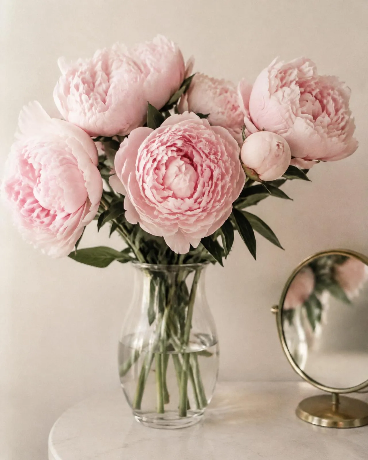 Blush peonies in a glass vase beside a small mirror